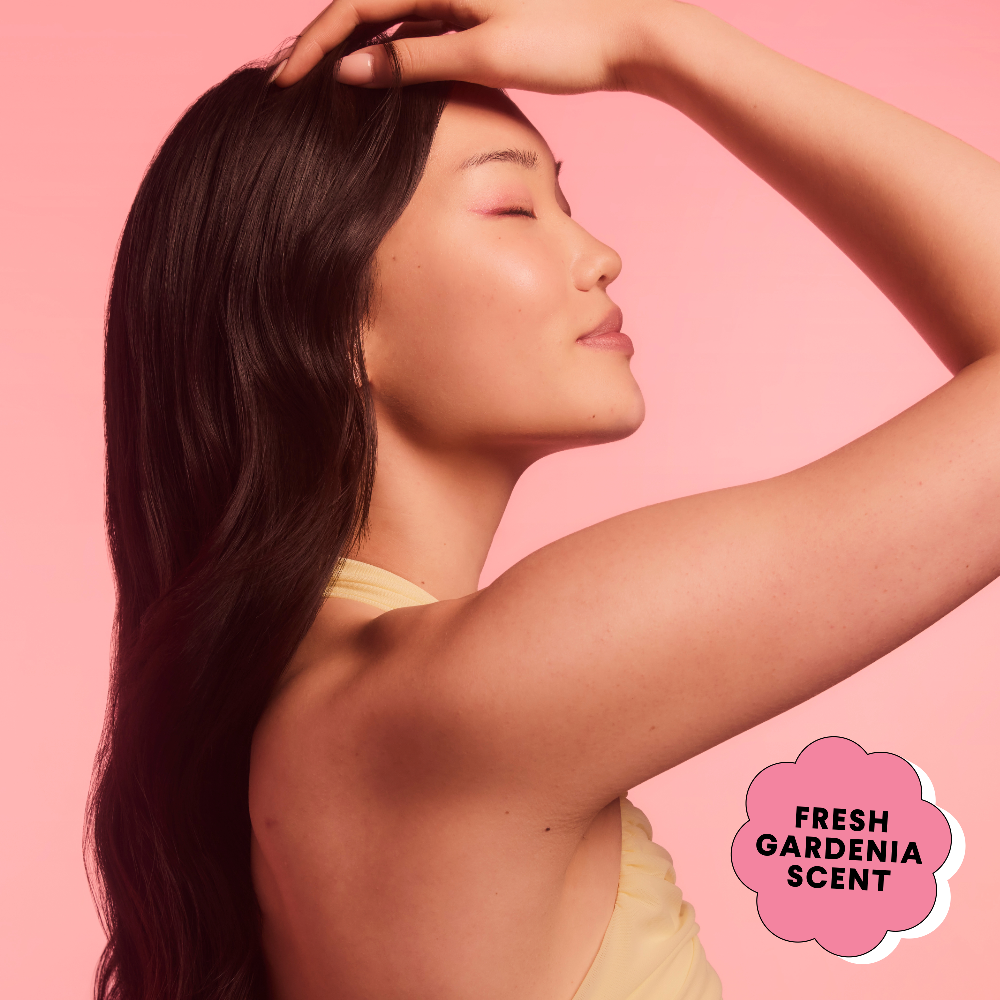 Woman runs her hand through her hair beside a Fresh Gardenia scent badge on pink.